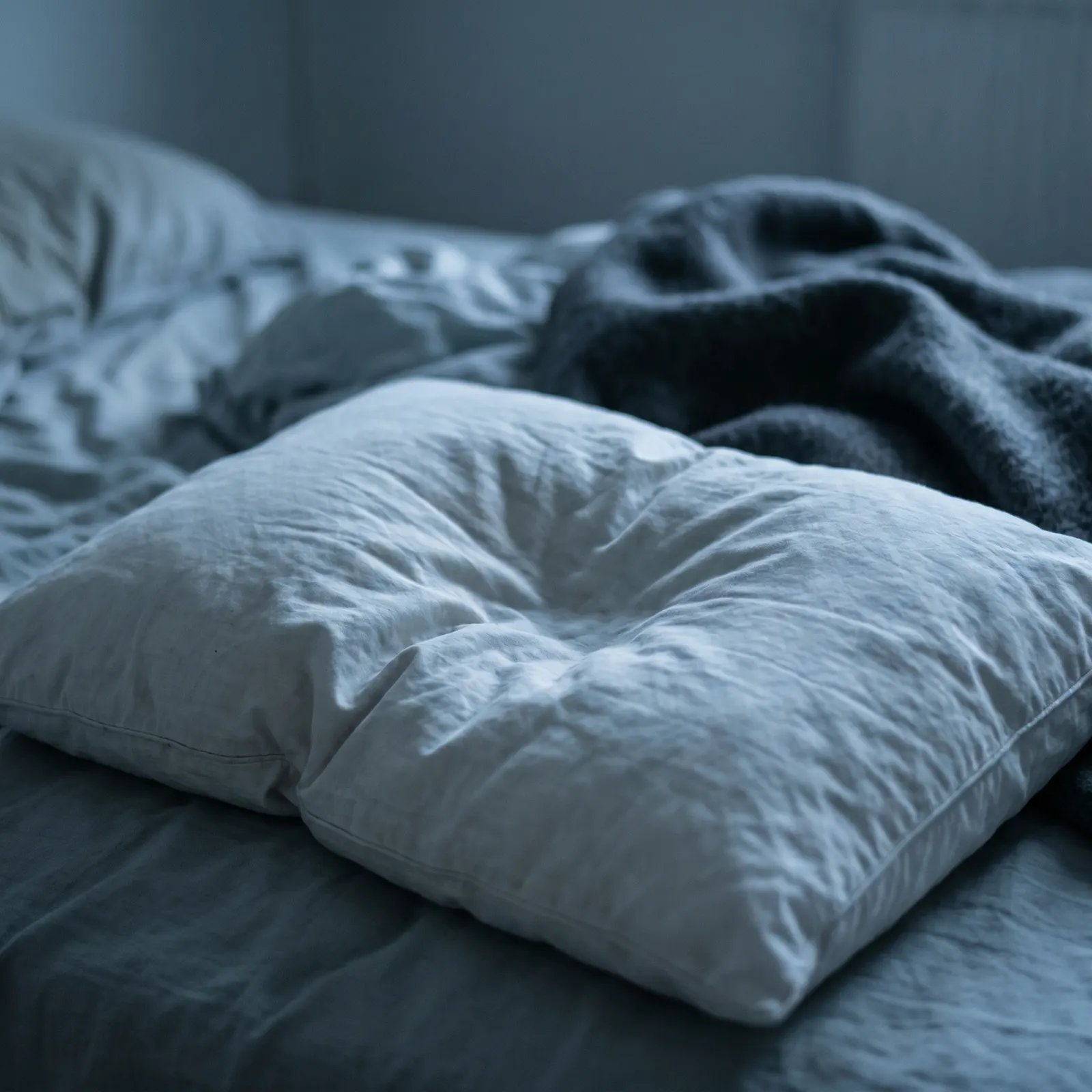 A tired, flattened pillow folded over on an unmade bed in cold morning light